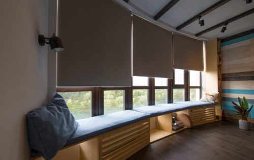 Motorized roller shades in the interior. Automatic roller blinds beige color on big glass windows. Remote Control Shades are above the windosill with pillows. Summer. Green trees outside.